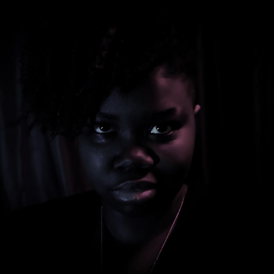 Dann a non-binary dark-skinned Afro-indigenous person stares into the camera intensely. Their dark hair swoops over their left eye and a necklace chain is visible around their neck. The picture is in muted sepia in tones of grays, browns and blacks.